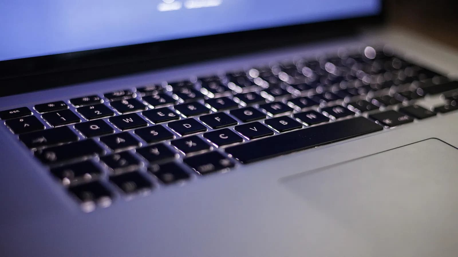 Close-up of a backlit laptop keyboard in low light, with a blurred screen glowing blue behind it