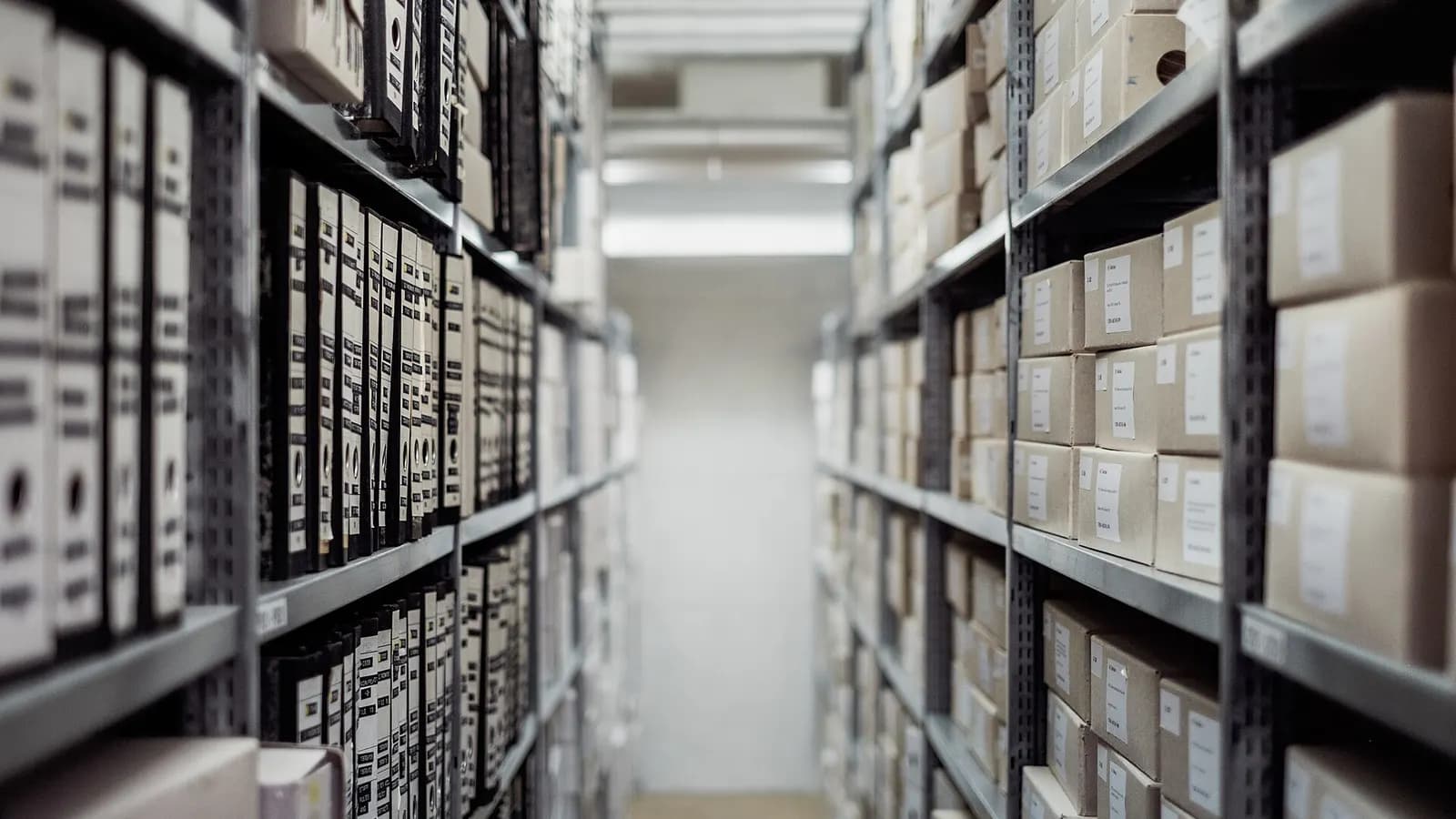 A narrow aisle between tall shelves of archive binders and boxes, with the far end softly out of focus