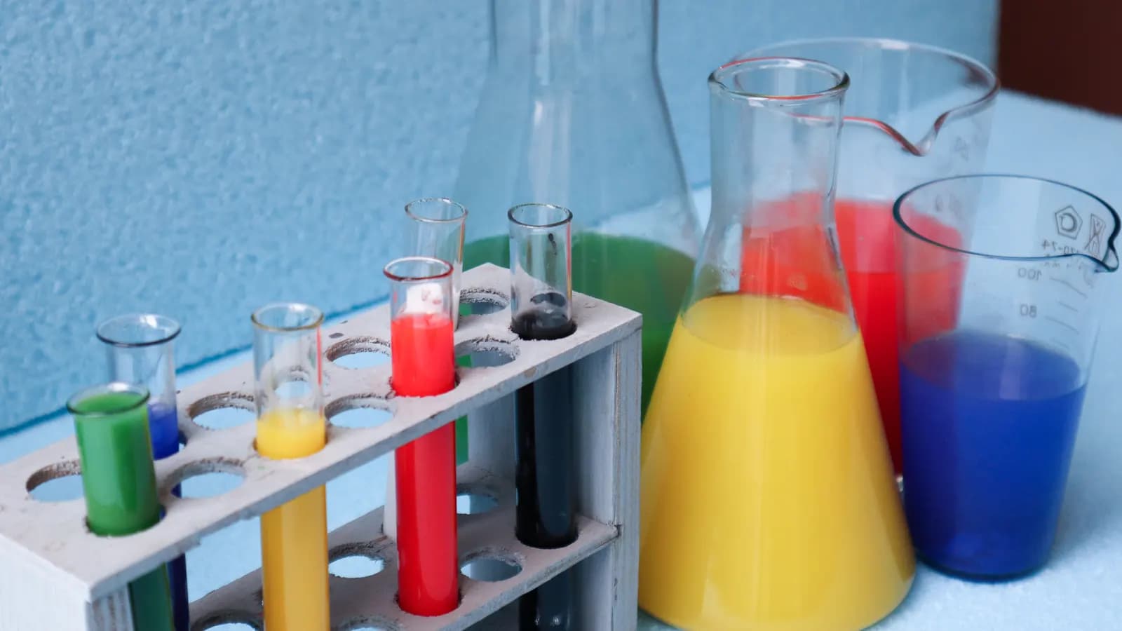 Test tubes in a rack and flasks filled with brightly coloured liquids on a pale blue background
